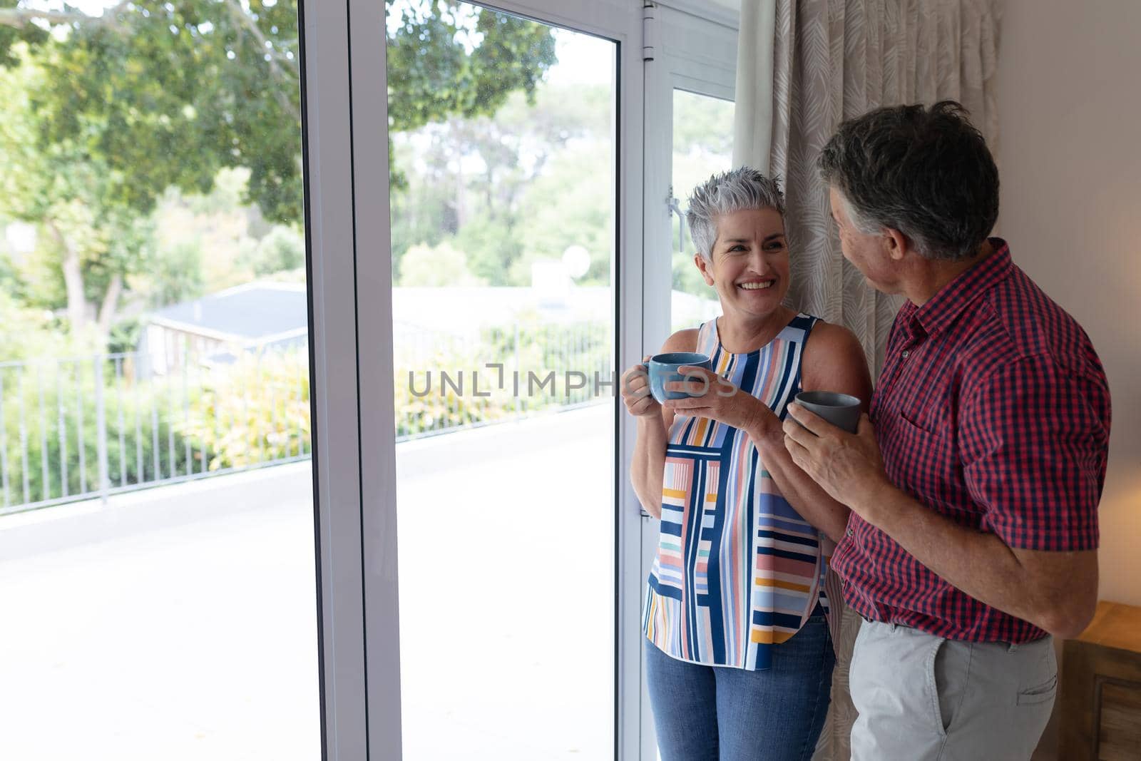 Happy senior caucasian couple standing by window talking holding cups of coffee. staying at home in isolation during quarantine lockdown.