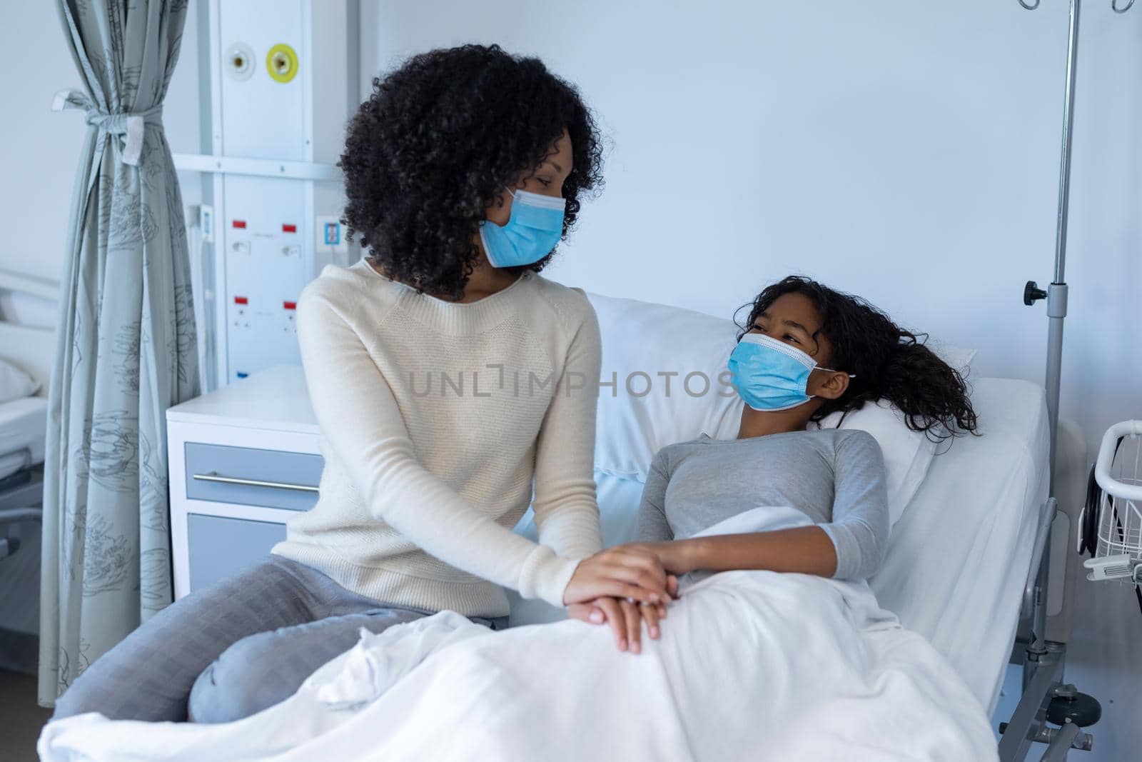 Mixed race mother comforting ill daughter in hospital bed, holding hands, both wearing face masks. medicine, health and healthcare services during coronavirus covid 19 pandemic.