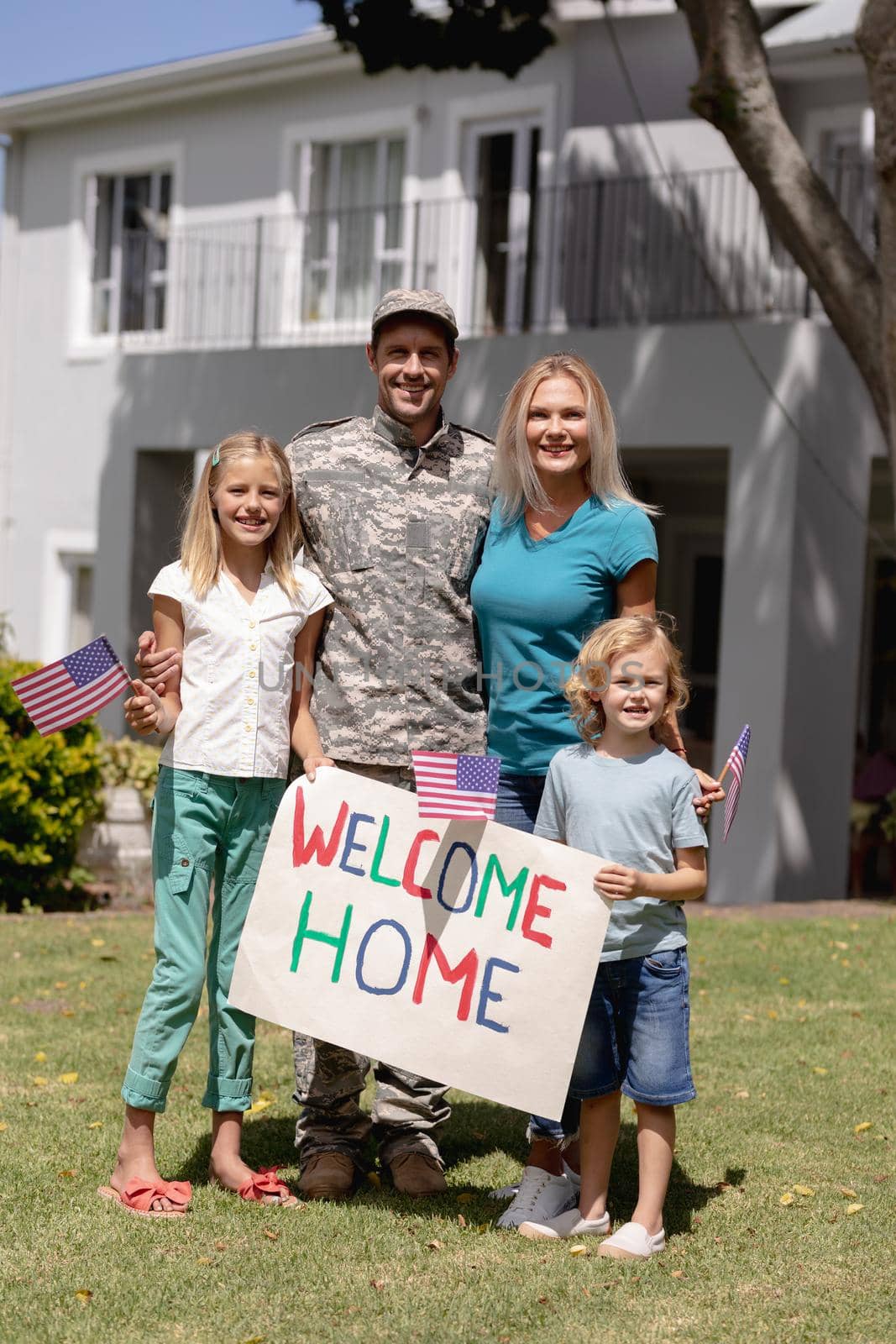 Happy caucasian soldier father embracing wife and children with welcome sign and flags outside home by Wavebreakmedia