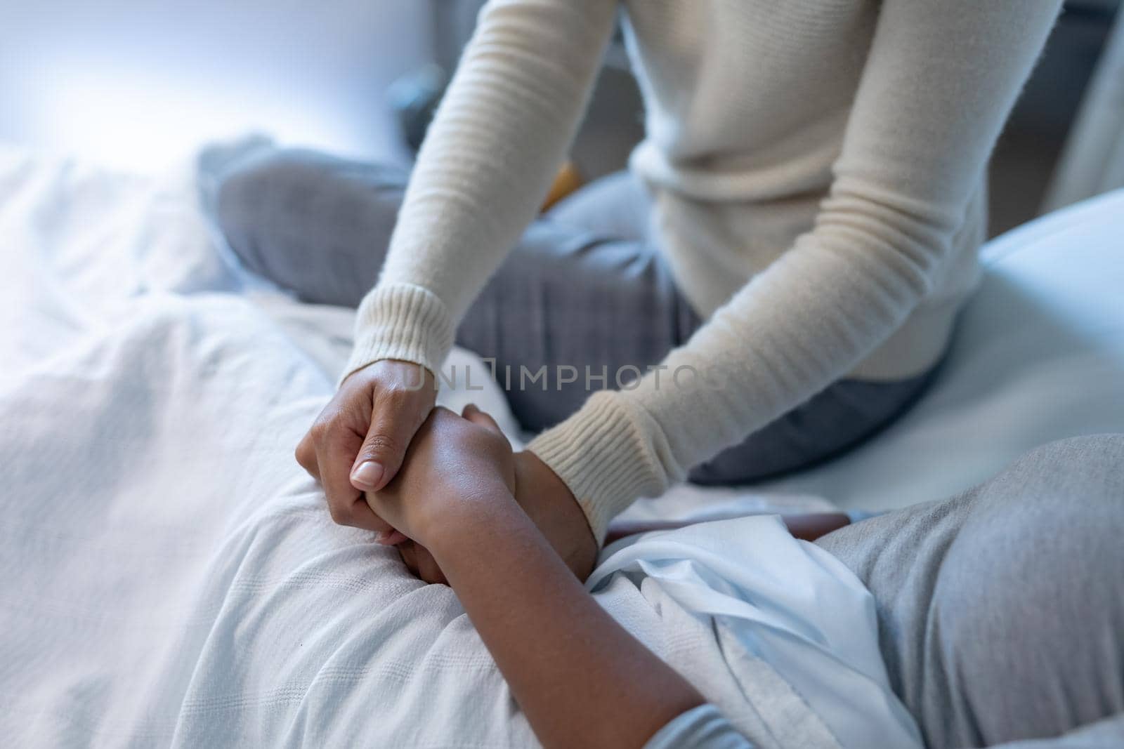 Midsection of mixed race mother comforting sick daughter, sitting on hospital bed holding her hands by Wavebreakmedia