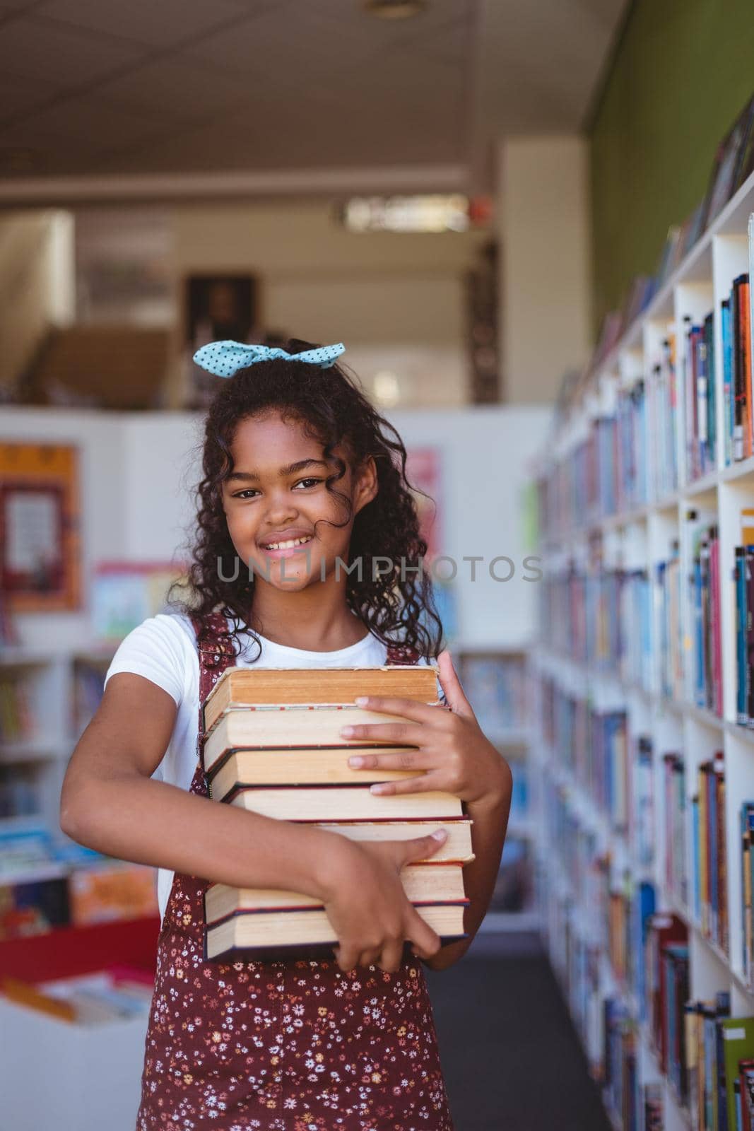 Portrait of smiling african american schoolgirl carrying stack of books in school library by Wavebreakmedia