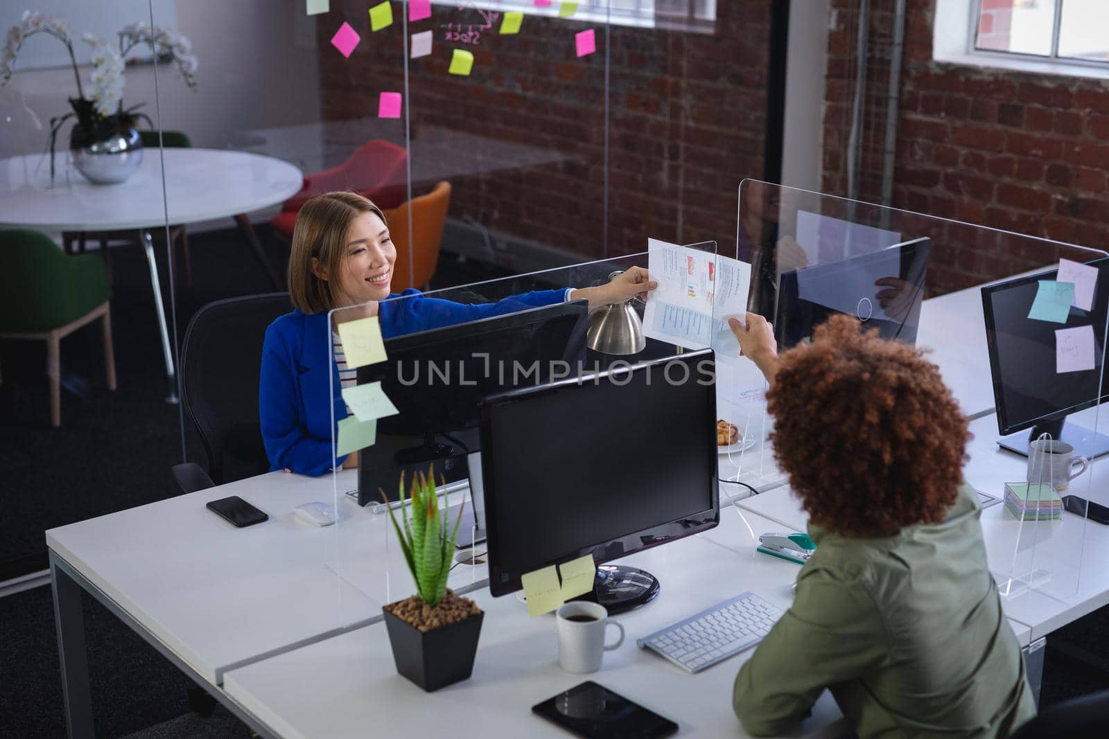 Diverse male and female colleagues in front of computers separated by sneeze shield giving document by Wavebreakmedia