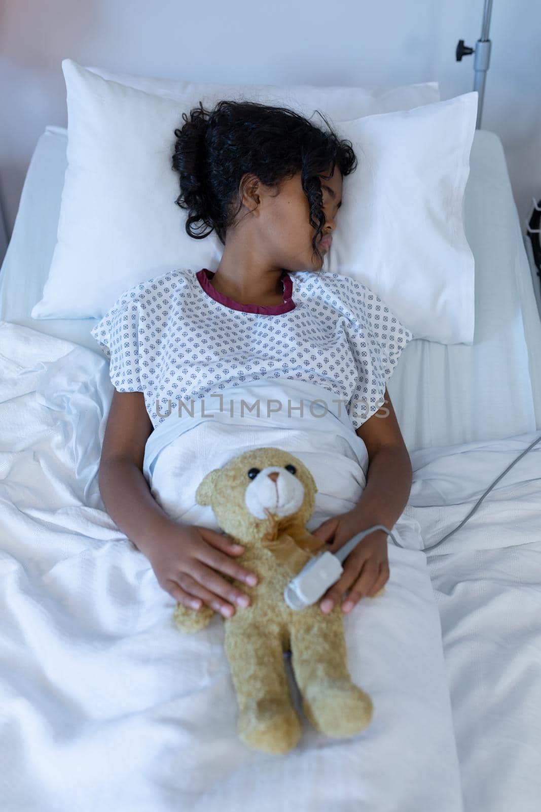 Sick mixed race girl asleep in hospital bed wearing fingertip pulse oximeter and holding teddy bear. medicine, health and healthcare services.
