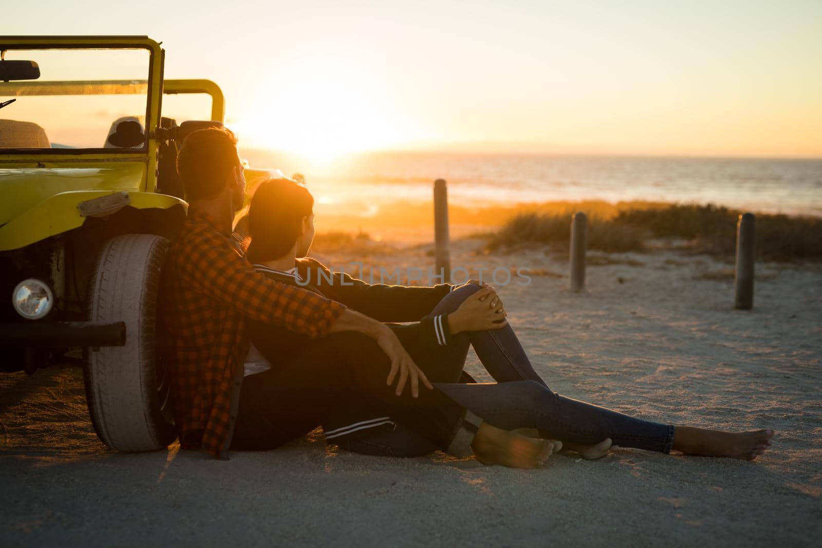 Happy caucasian couple leaning against beach buggy by the sea watching sunset. beach break on summer holiday road trip.