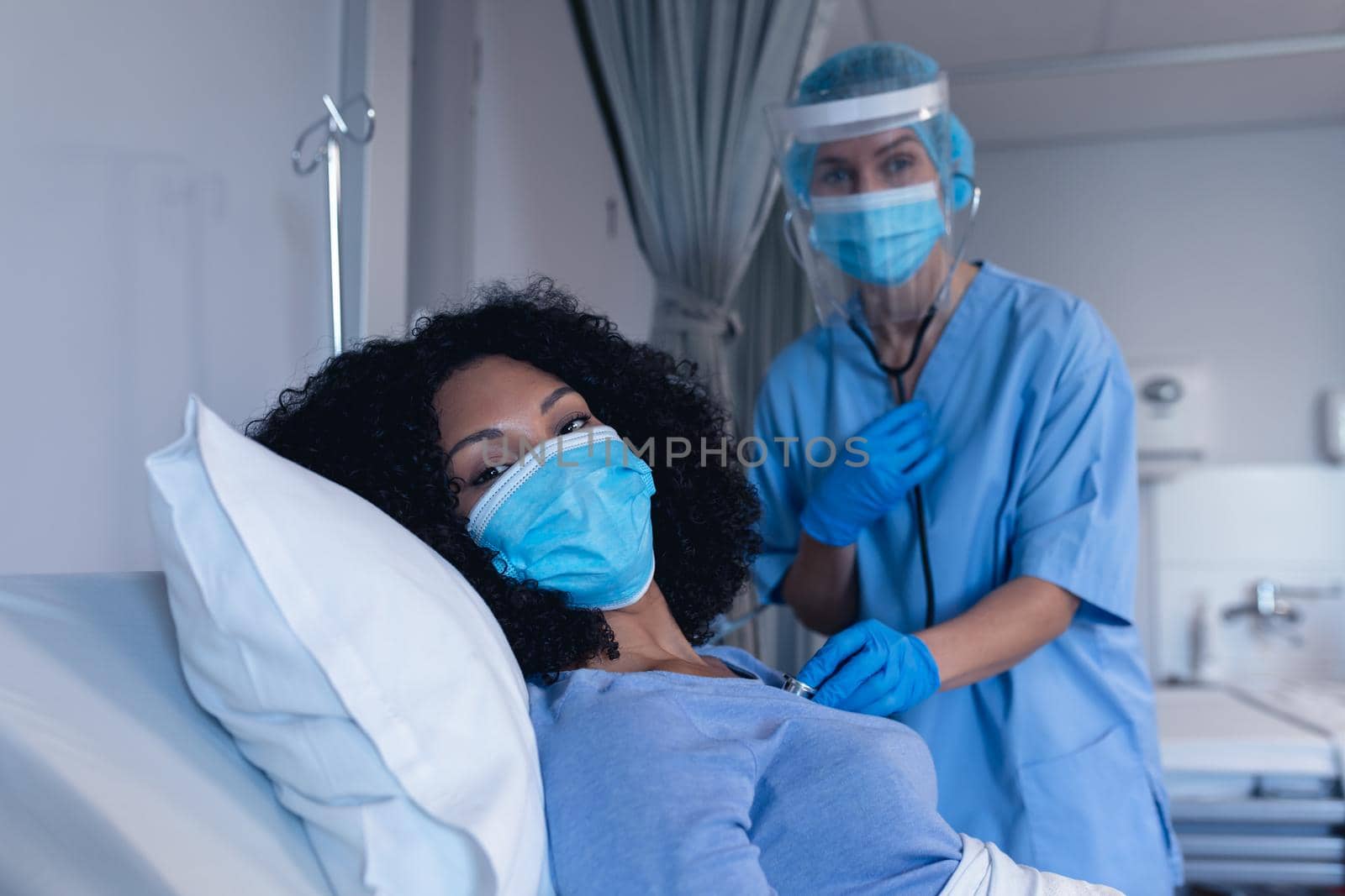 Caucasian female doctor in hospital in face mask examining female patient with stethoscope by Wavebreakmedia