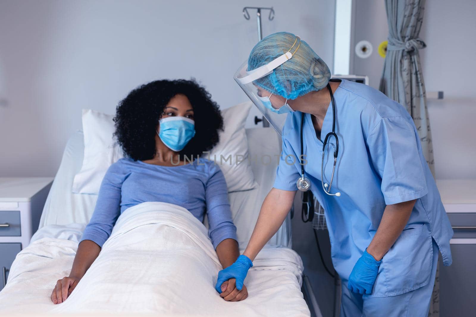Caucasian female doctor in hospital in face mask consoling female patient in bed wearing face mask by Wavebreakmedia