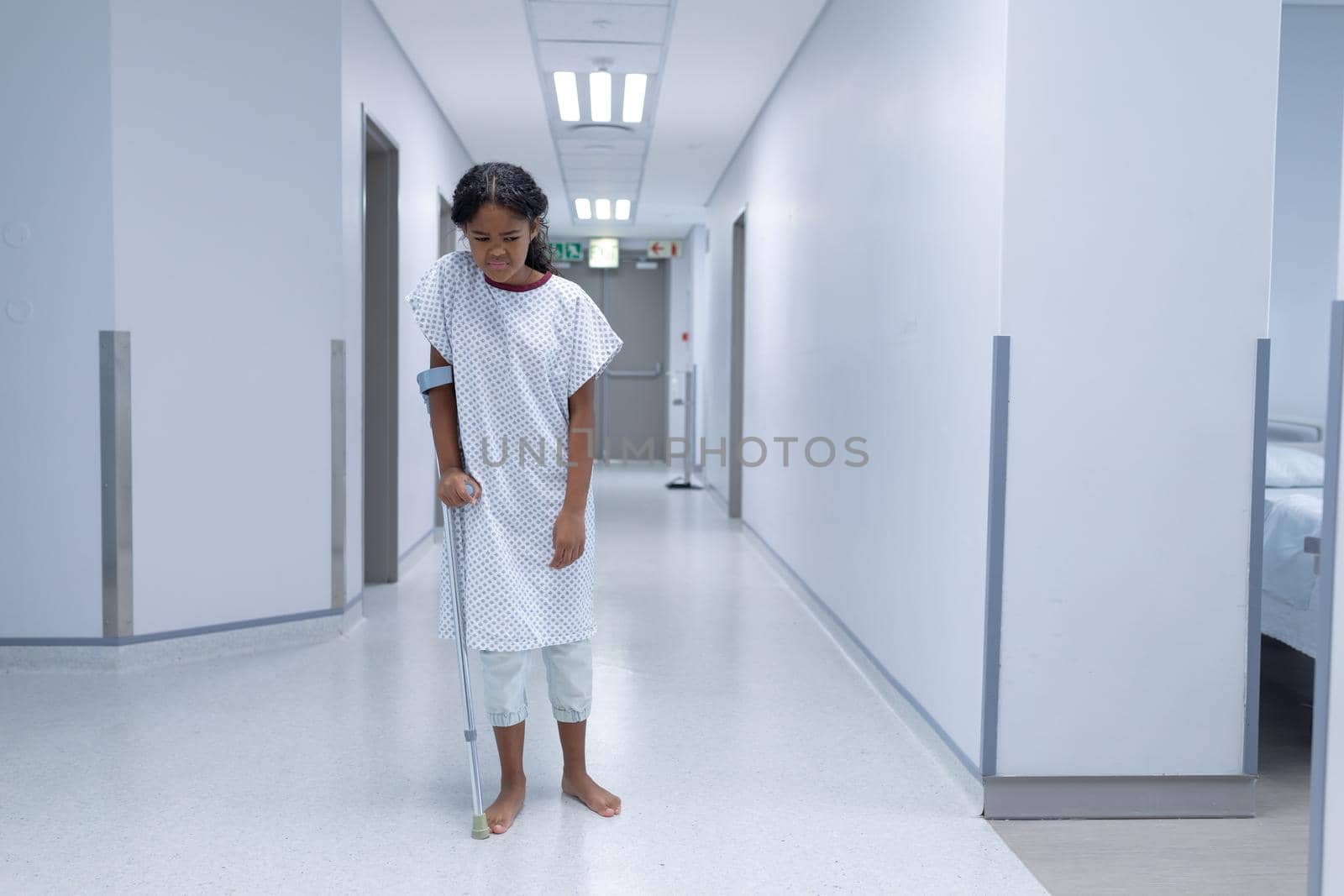 Sick mixed race girl walking barefoot in hospital corridor using a crutch. medicine, health and healthcare services.