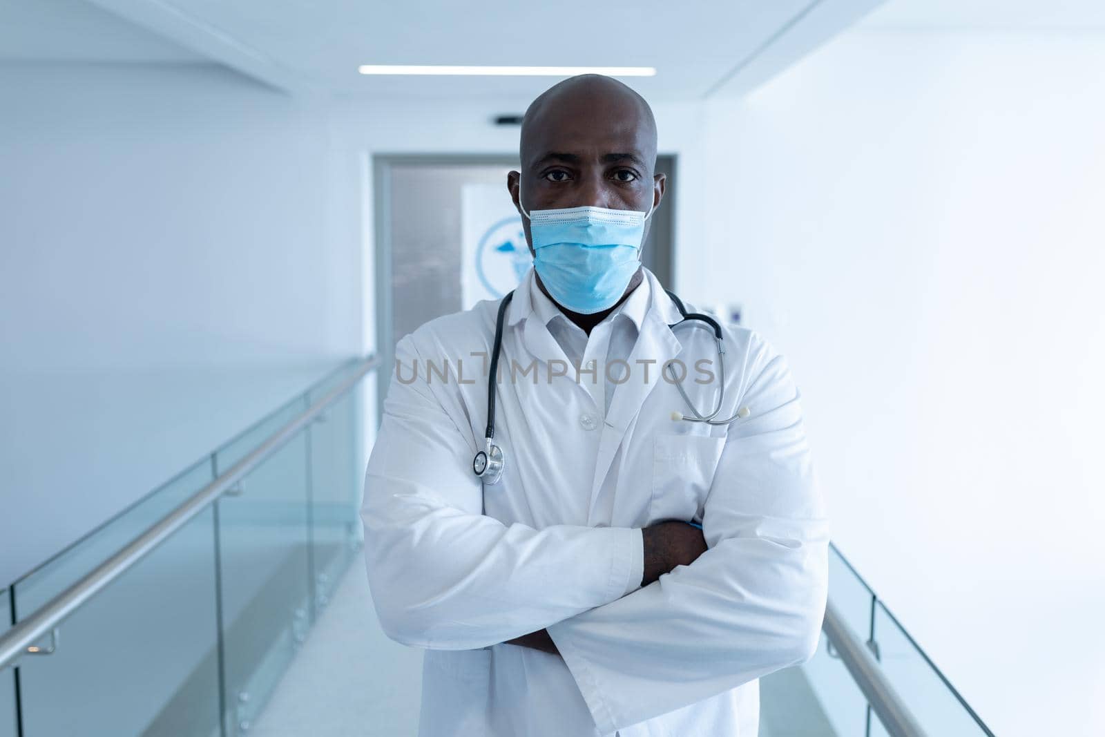 Portrait of african american male doctor in hospital corridor with arms crossed wearing face mask. medical professional at work during coronavirus covid 19 pandemic.