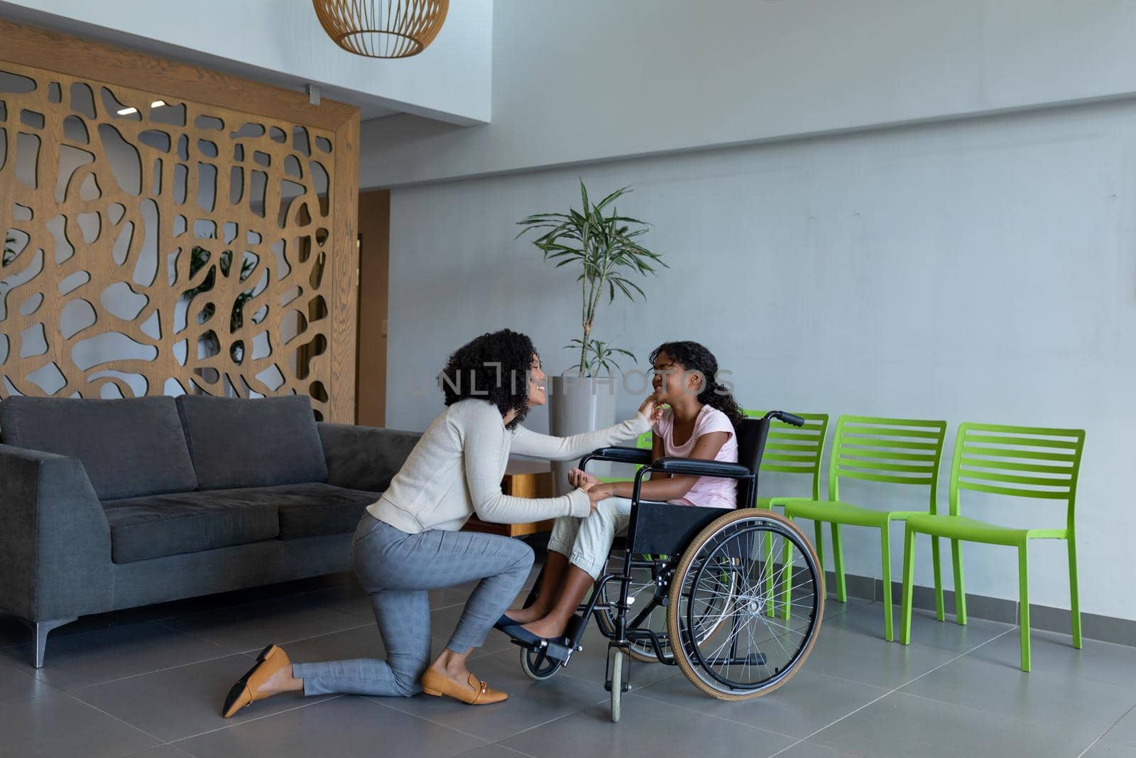 Happy mixed race mother kneeling holding hand of smiling daughter in wheelchair in hospital foyer. medicine, health and healthcare services.