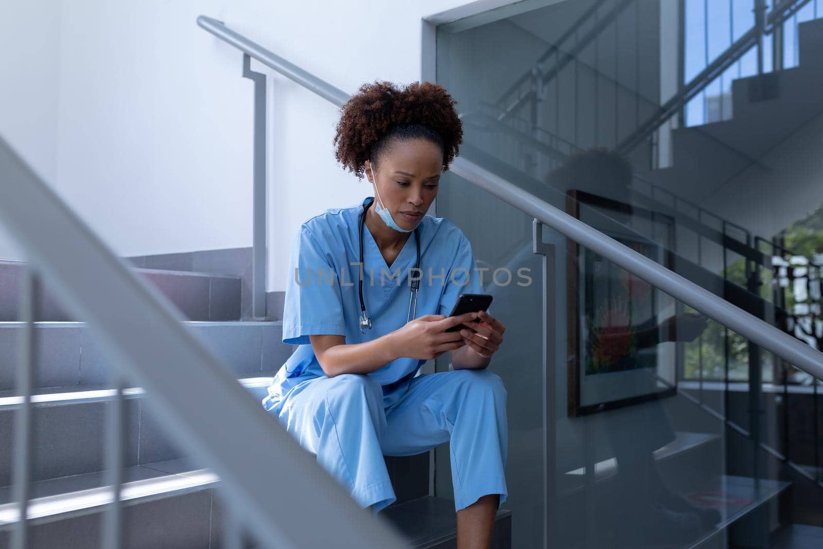 Mixed race female doctor with face mask using smartphone sitting on stairs in hospital. medical professional at work during coronavirus covid 19 pandemic.