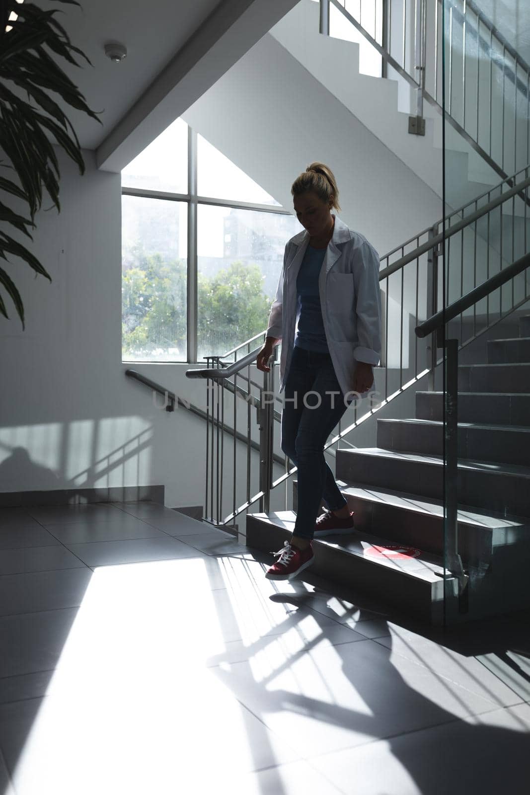 Caucasian female doctor going downstairs on hospital staircase during sunny day. medicine, health and healthcare services.