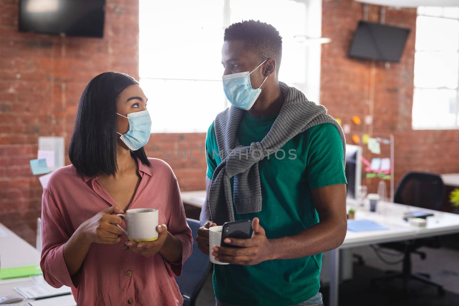 Diverse business colleagues wearing face masks having coffee in office. independent creative design business during covid 19 coronavirus pandemic.