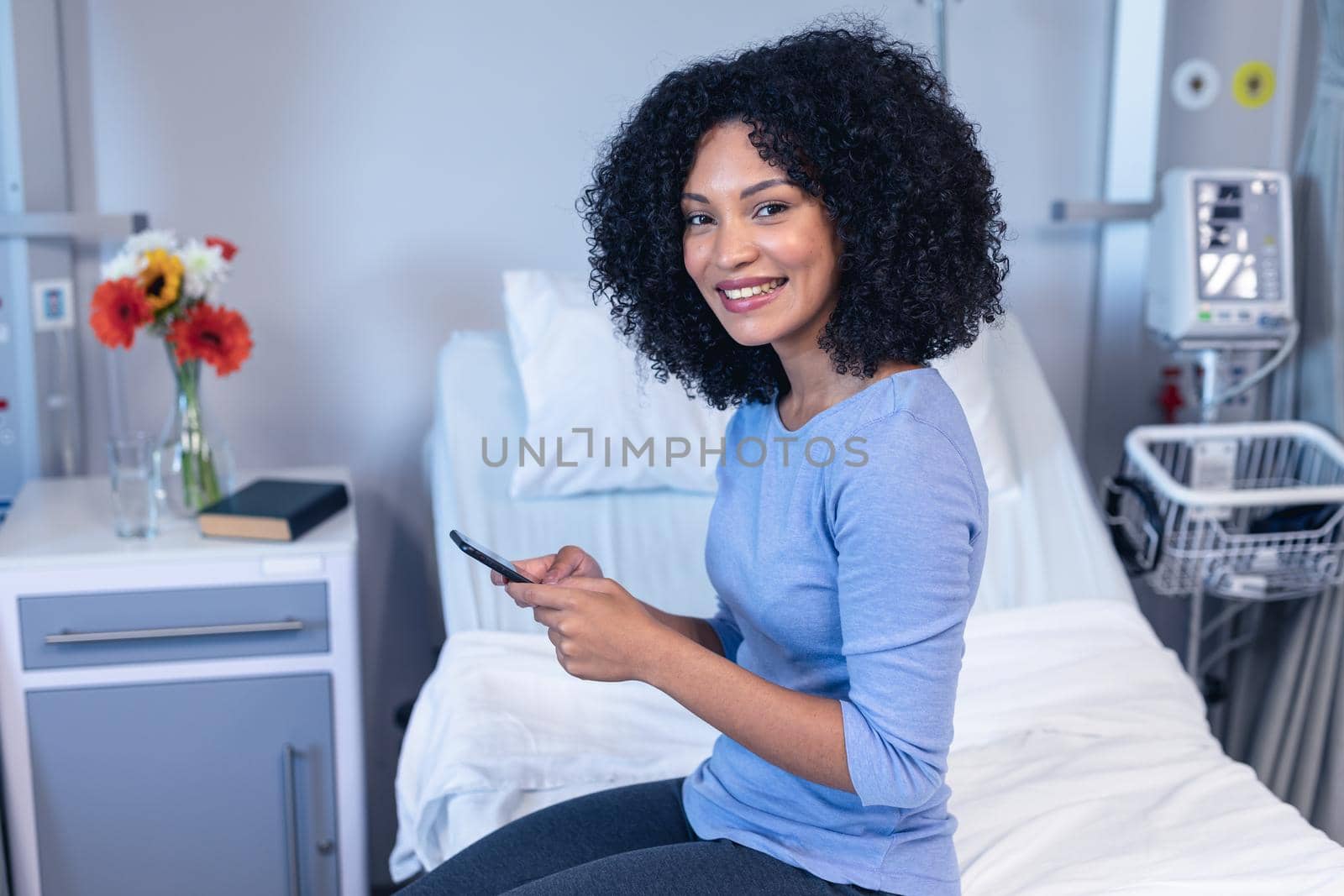 Smiling mixed race female patient sitting on hospital bed using smartphone and looking to camera. medicine, health and healthcare services.