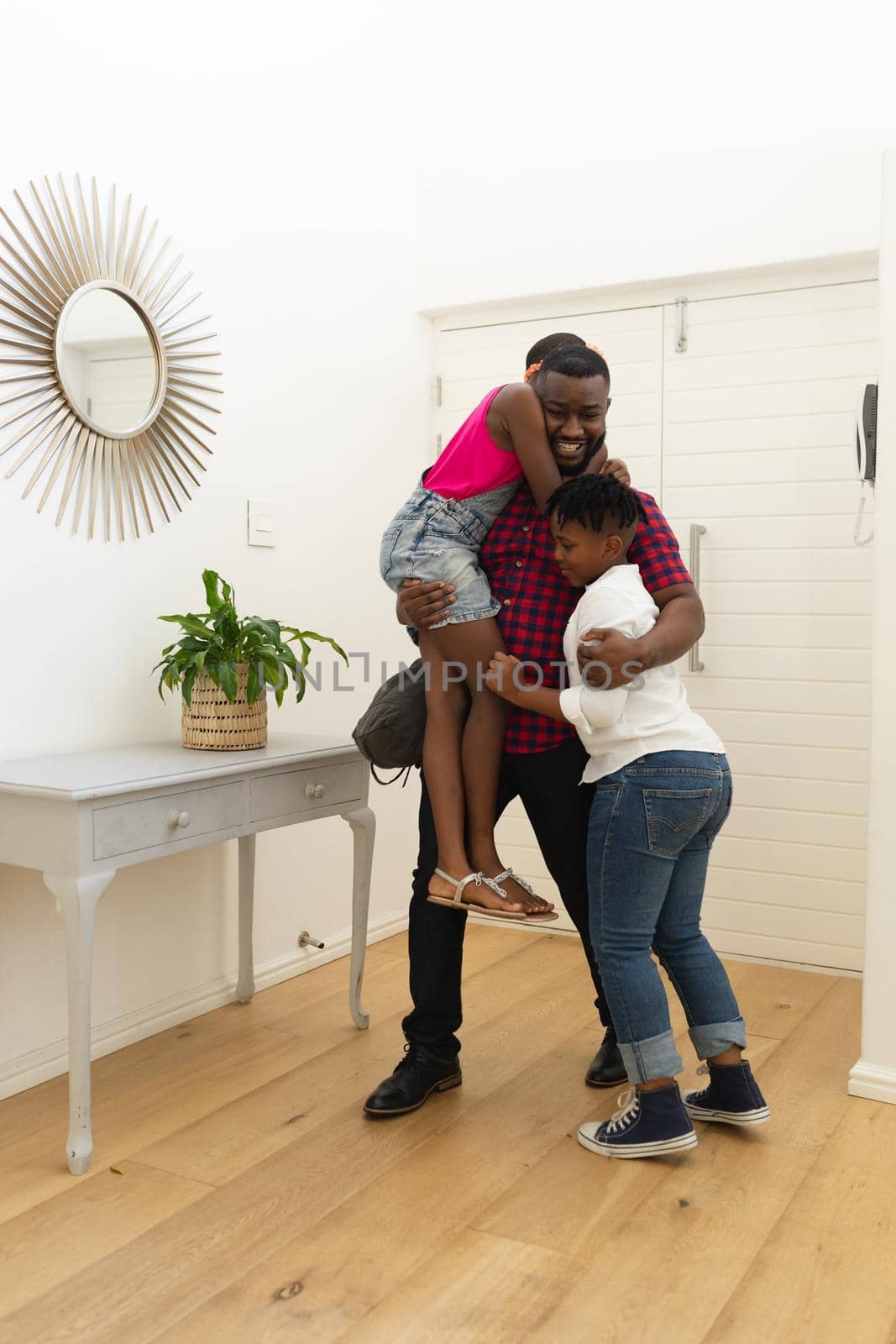 Smiling african american father returning home embracing son and daughter in hallway. happy family at home together.