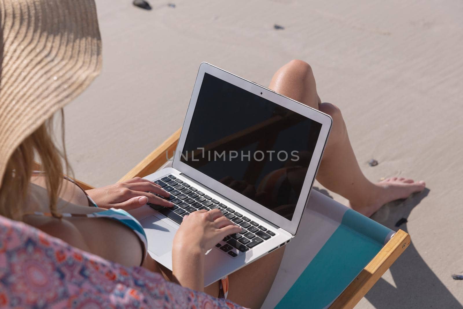 Mid section of caucasian woman using laptop while sitting on deck chair at the beach. summer beach holiday concept.