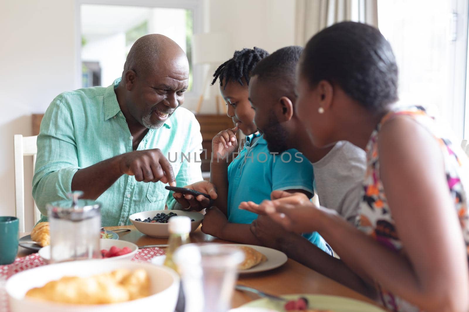 African american grandfather using smartphone with grandson and his parents at table. three generation family spending quality time together.