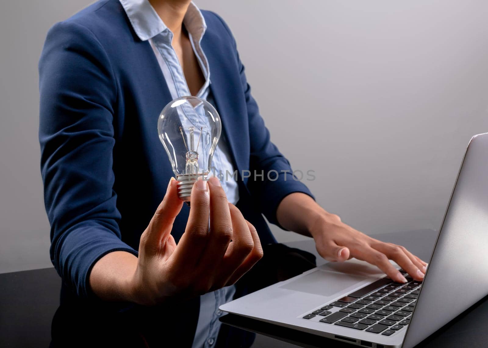 Mid section of businesswoman holding a light bulb using laptop against grey background. business, professionalism and technology concept