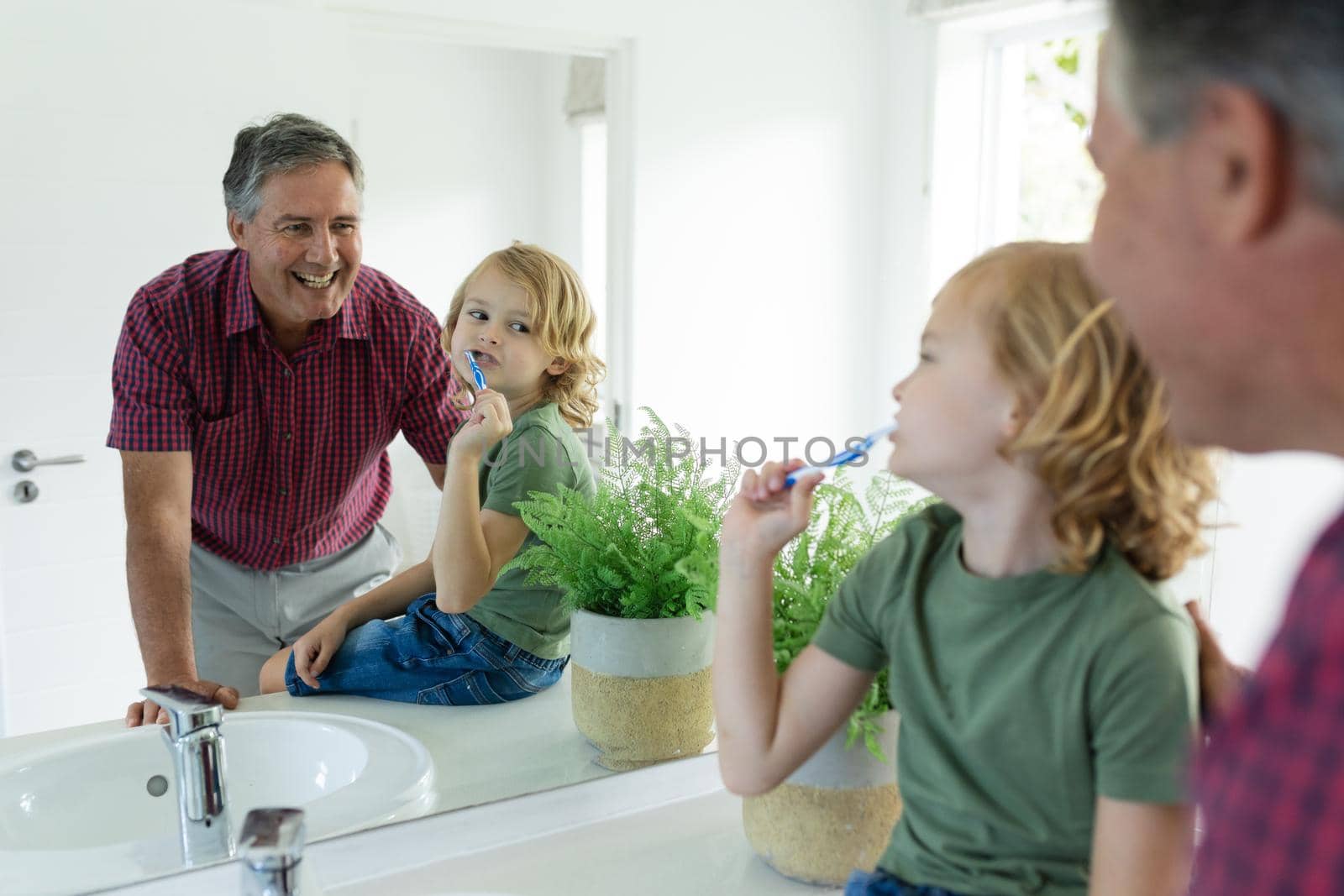 Smiling caucasian grandfather in bathroom with grandson brushing teeth both looking in mirror. staying at home in isolation during quarantine lockdown.
