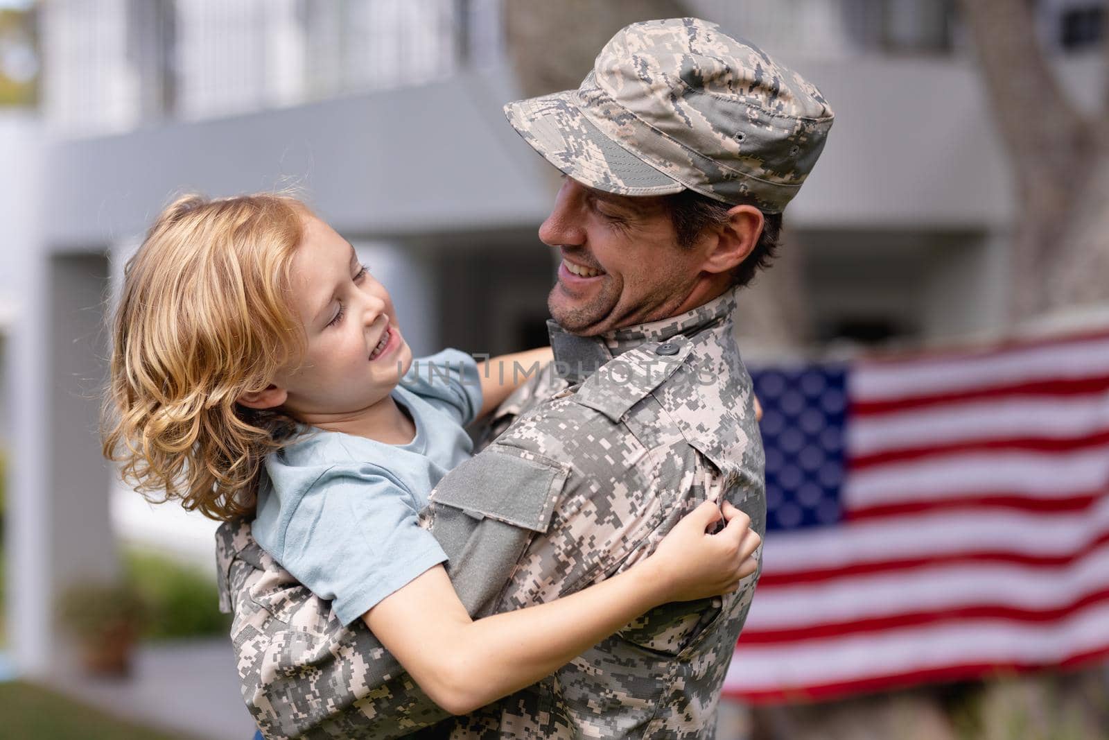 Smiling caucasian soldier father carrying son in garden with american flag hanging outside house by Wavebreakmedia