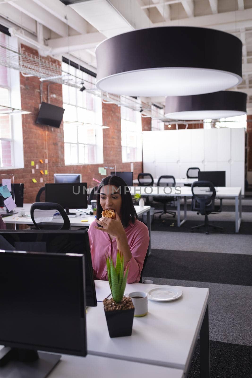Mixed race businesswoman sitting in office in front of computer and having snack. independent creative design business.