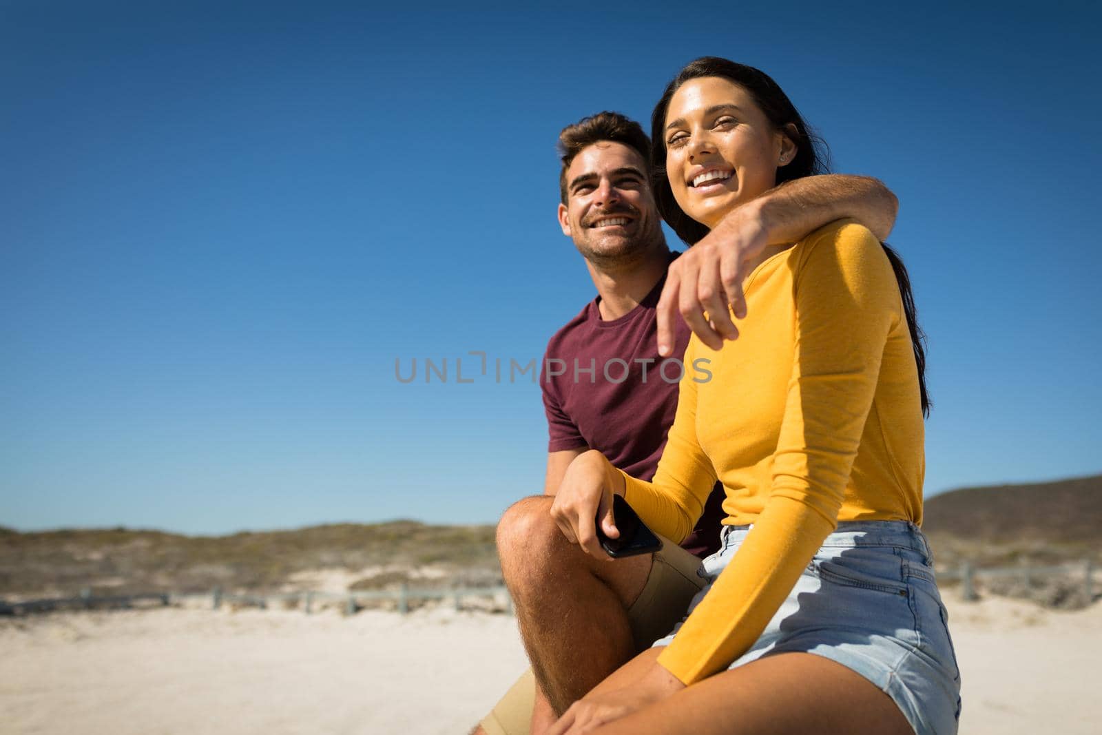 Happy caucasian couple on the beach embracing woman holding smartphone. vacation outdoor leisure time by the sea.
