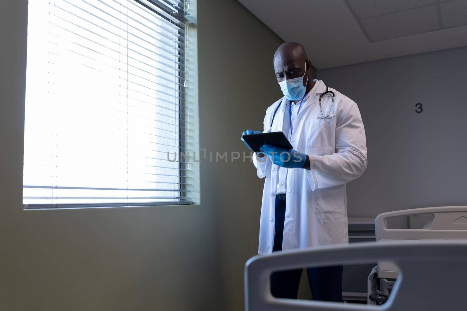 African american male doctor in hospital wearing face mask using tablet. medical professional at work during coronavirus covid 19 pandemic.