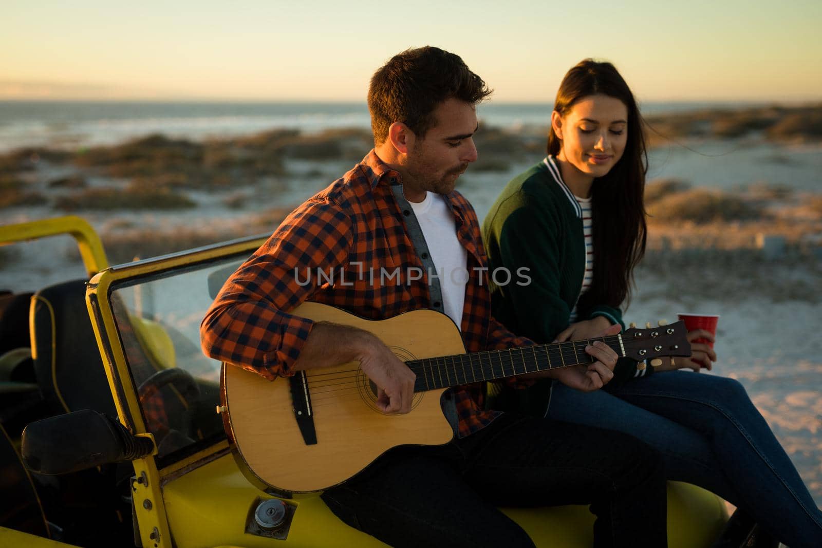 Happy caucasian couple sitting on beach buggy by the sea playing guitar and drinking during sunset. beach break on summer holiday road trip.