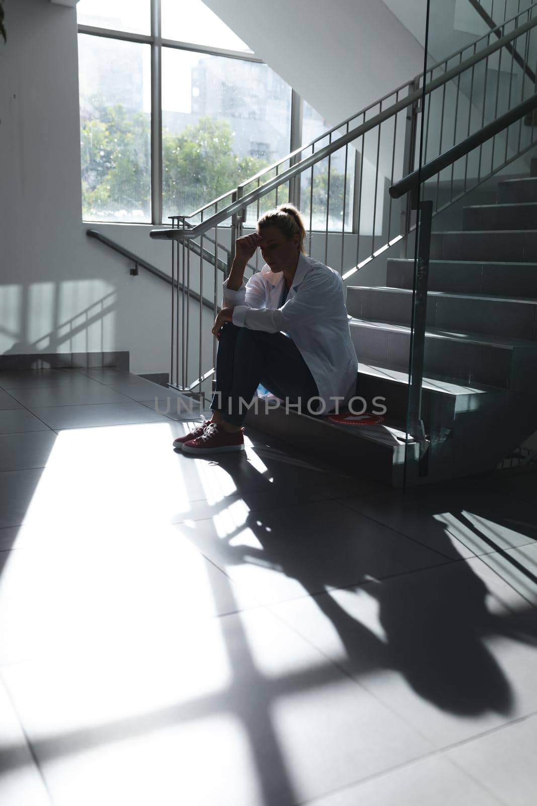 Caucasian female doctor sitting on sunny hospital staircase and worrying by Wavebreakmedia