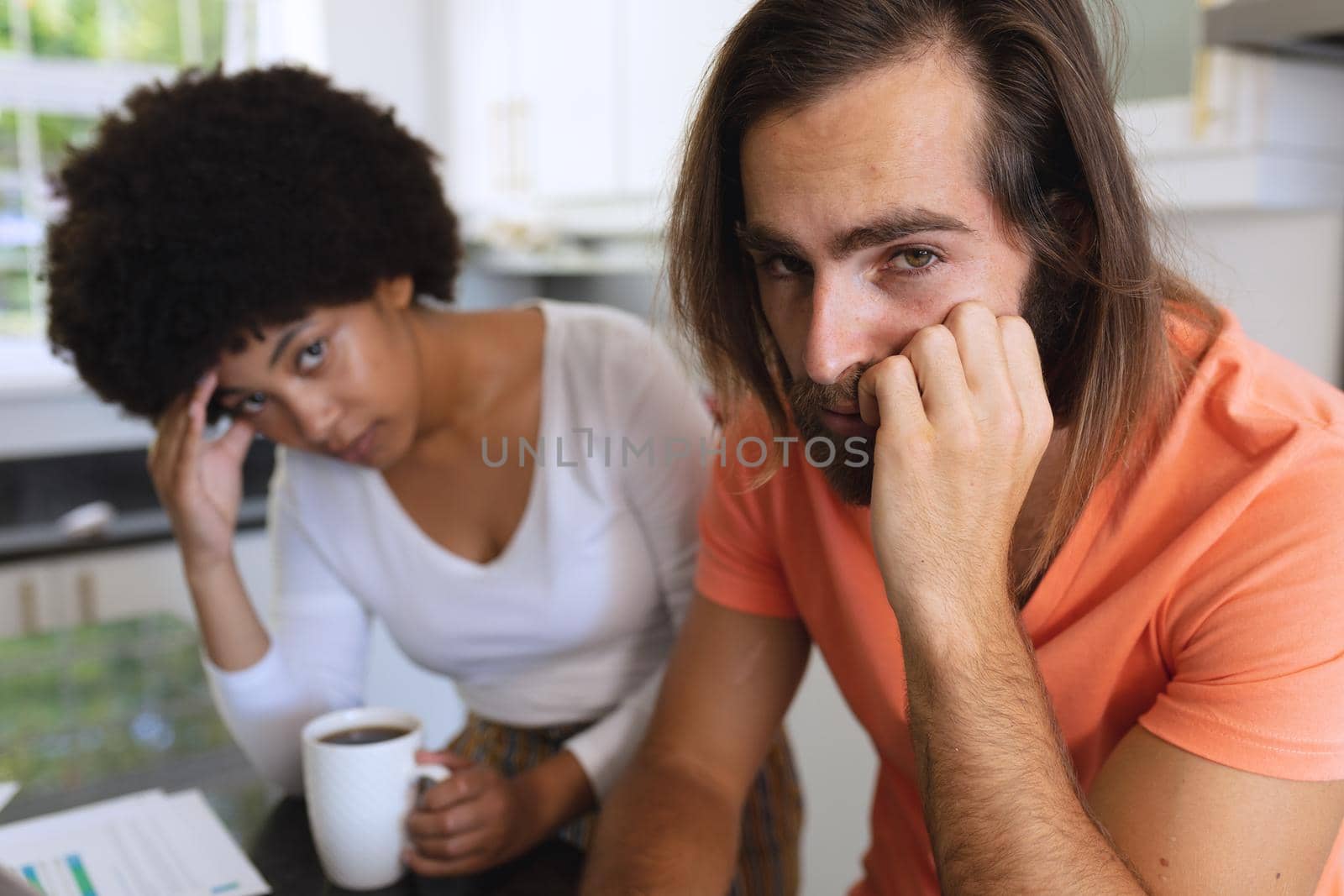 Portrait of worried diverse couple sitting in kitchen looking at camera. staying at home in isolation during quarantine lockdown.