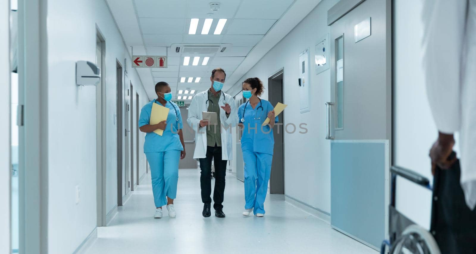Diverse group of male and female doctors wearing face masks walking looking at tablet. medicine, health and healthcare services during coronavirus covid 19 pandemic.