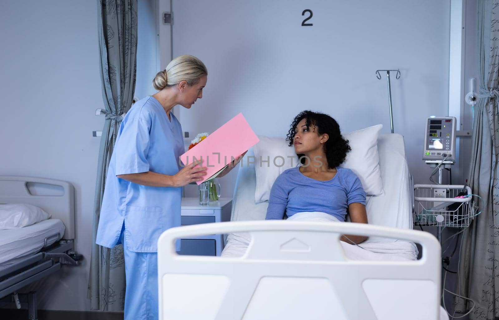 Caucasian female doctor holding file talking with mixed race patient sitting up in hospital bed by Wavebreakmedia