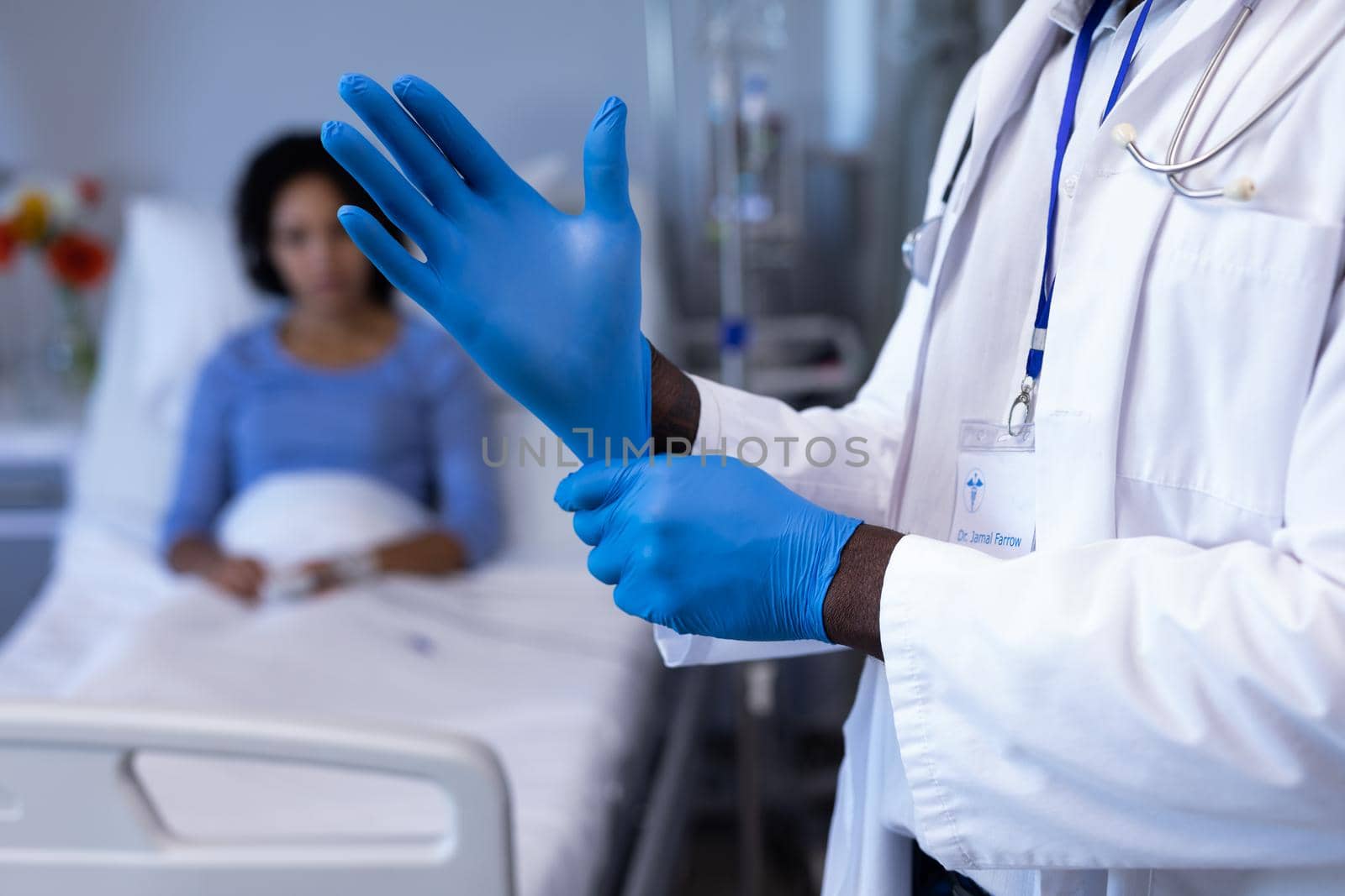 Midsection of african american male doctor putting on gloves, woman in hospital bed in background. medicine, health and healthcare services during coronavirus covid 19 pandemic.