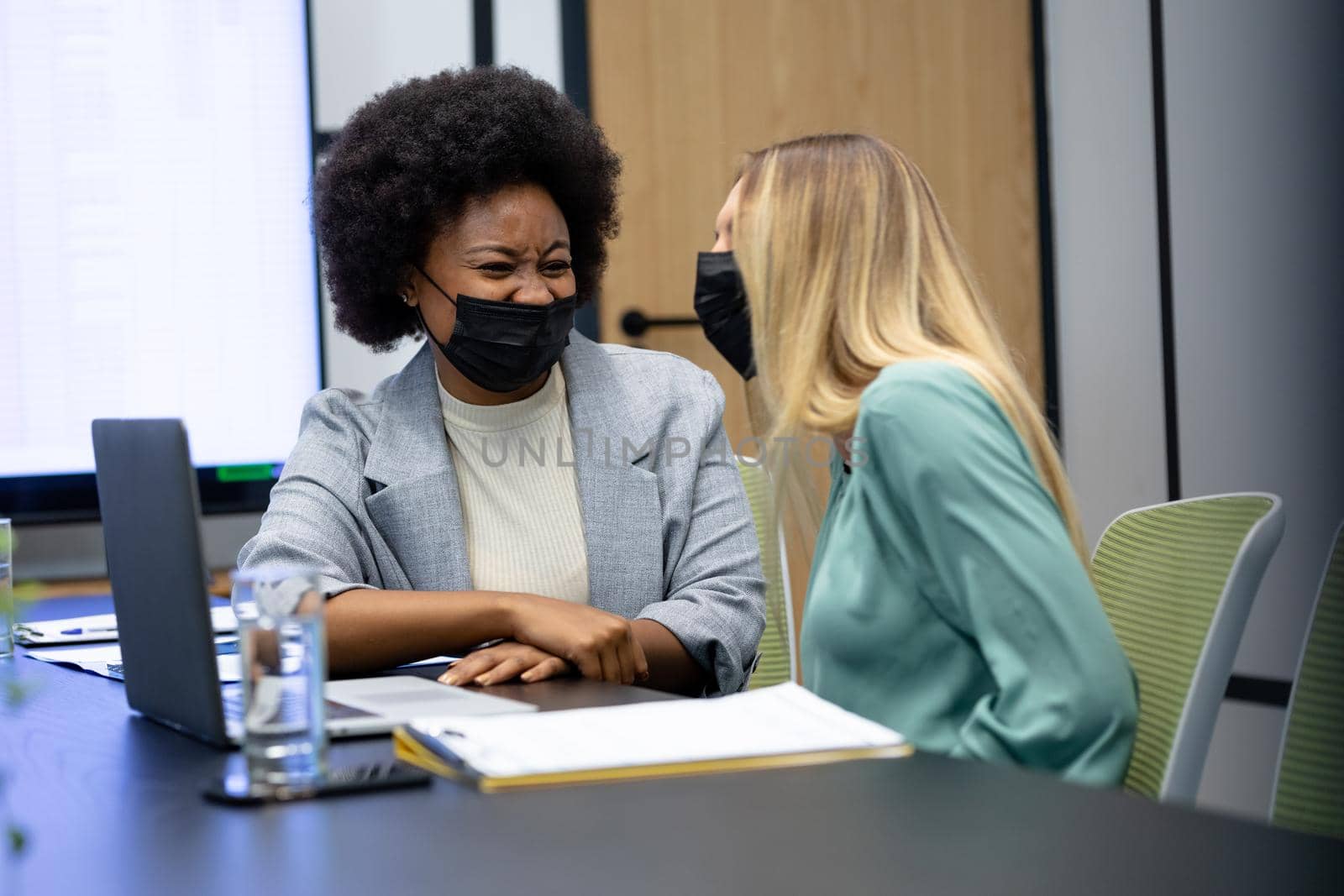 Diverse female business colleagues wearing masks laughing in meeting room by Wavebreakmedia