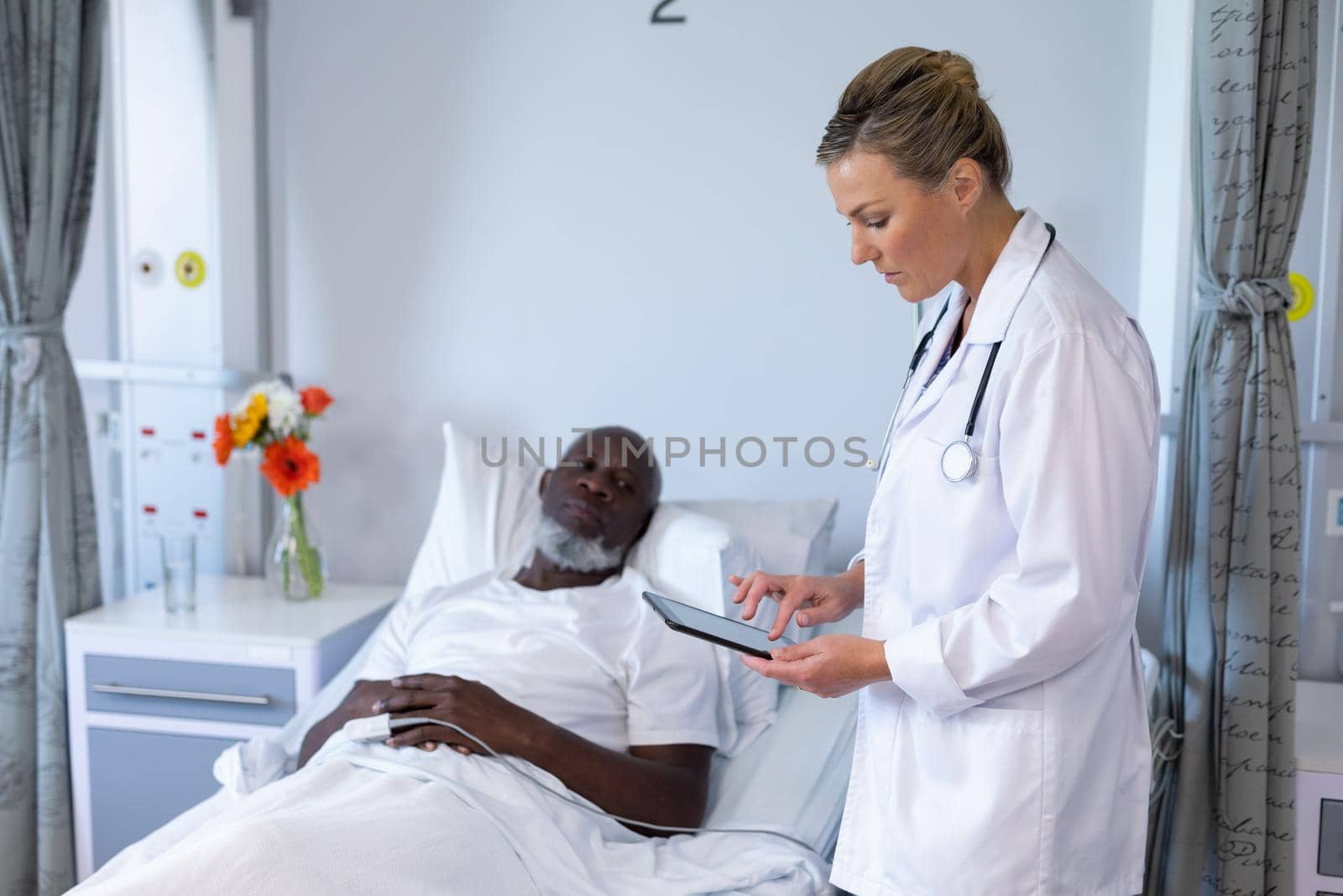 Caucasian female doctor standing next to african american male in hospital patient room using tablet. medicine, health and healthcare services.
