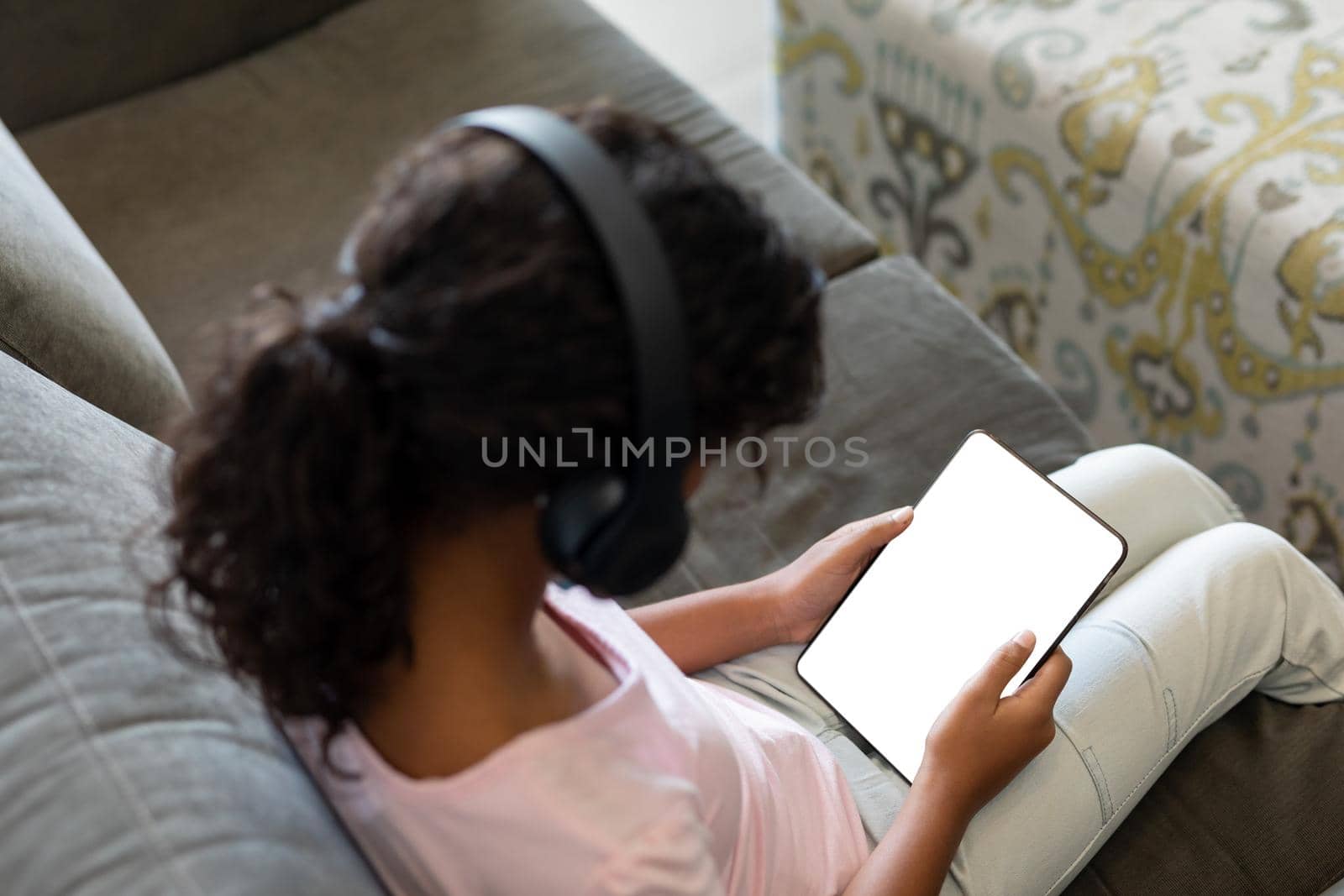 Elevated view of mixed race girl wearing wireless headphones sitting on couch using digital tablet. teenage lifestyle, leisure time, communication and technology.