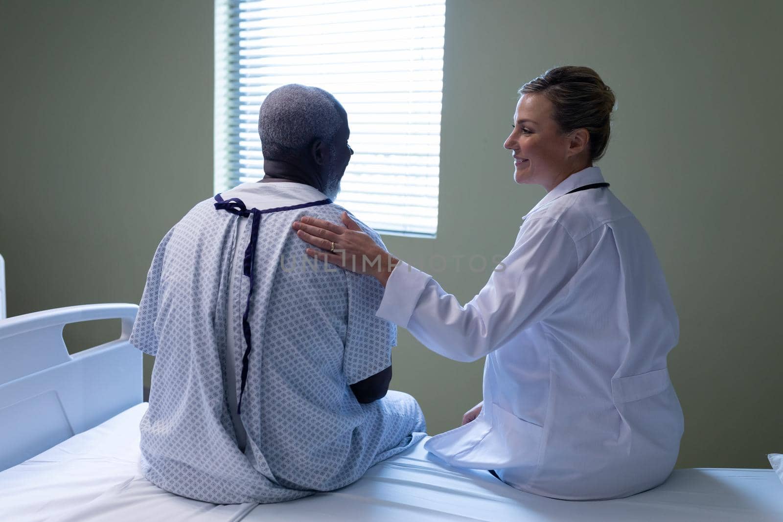 Diverse female doctor and male patient sitting on hospital bed smiling to each other. medicine, health and healthcare services.