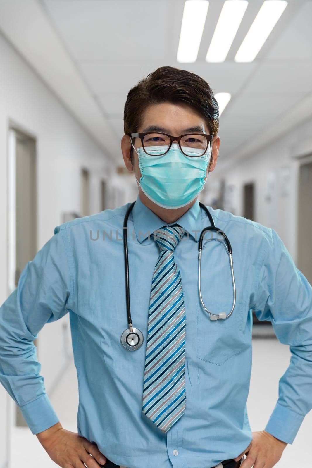 Portrait of mixed race male doctor wearing face mask standing in hospital corridor. medicine, health and healthcare services during coronavirus covid 19 pandemic.