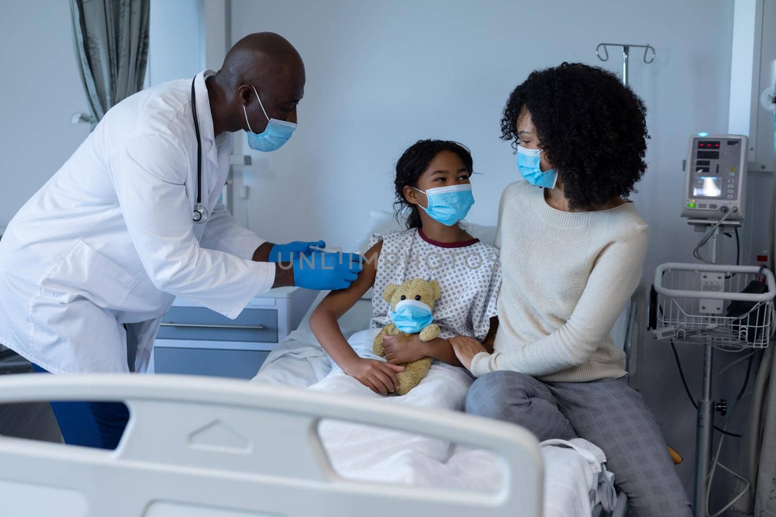 Male doctor giving covid vaccination to sick girl in hospital bed with mother, all in face masks. medicine, health and healthcare services during coronavirus covid 19 pandemic.