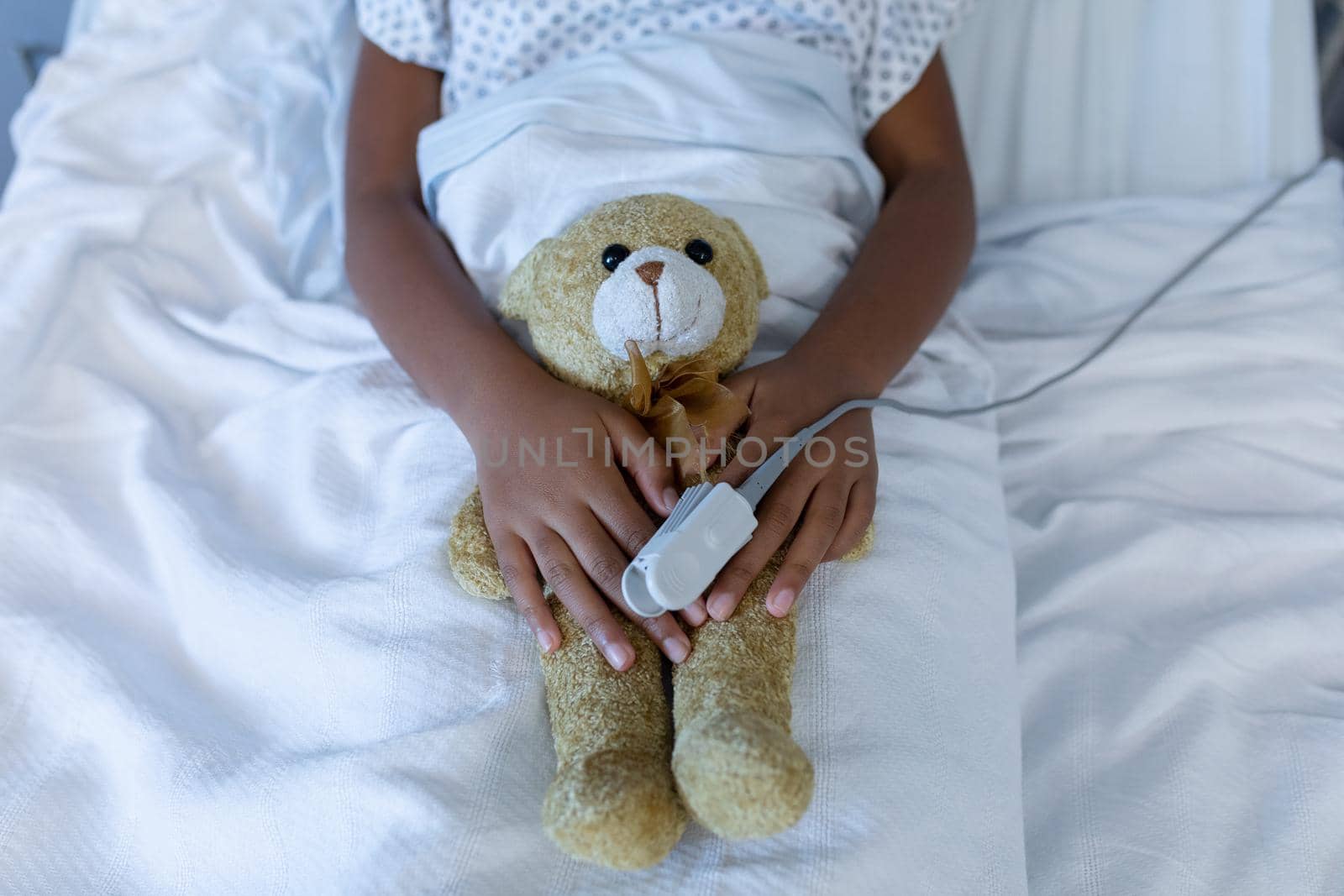 Midsection of sick mixed race girl in hospital bed wearing fingertip pulse oximeter holding teddy. medicine, health and healthcare services.