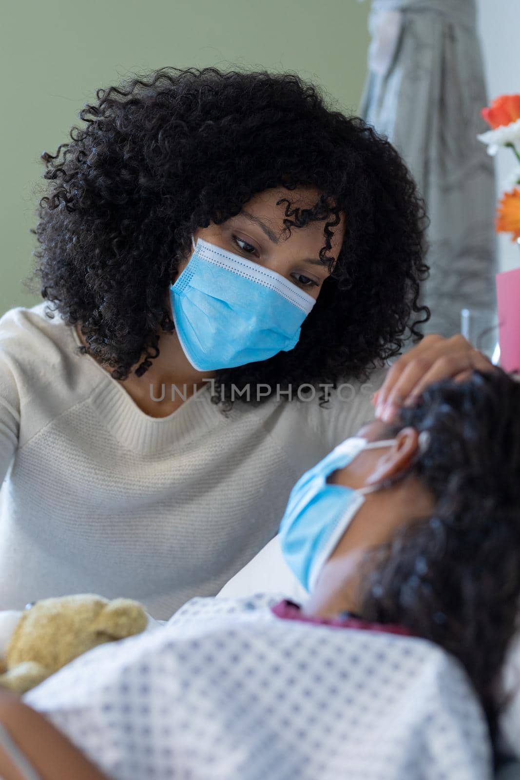 Mixed race mother and sick daughter in face masks in hospital, girl holding teddy bear. medicine, health and healthcare services during covid 19 coronavirus pandemic.