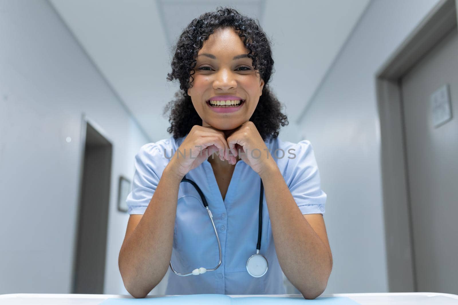 Mixed race female doctor at desk leaning chin on hands and smiling during video call consultation. telemedicine, online healthcare services during quarantine lockdown.