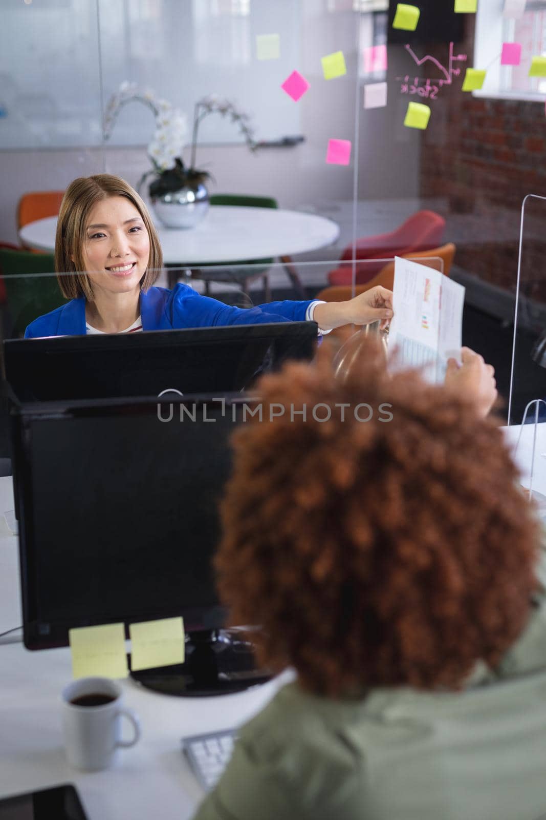 Diverse male and female colleagues in front of computers separated by sneeze shield giving document by Wavebreakmedia