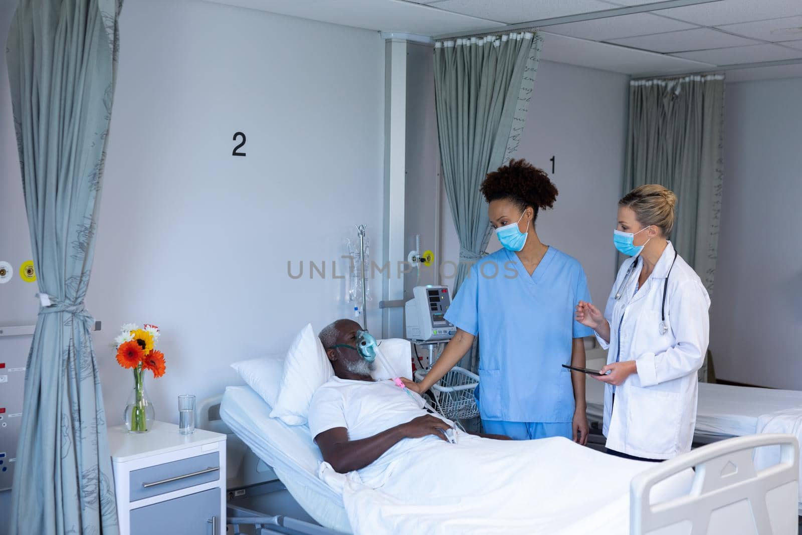 Two diverse female doctors wearing masks standing next to male patient in hospital room. medicine, health and healthcare services during coronavirus covid 19 pandemic.