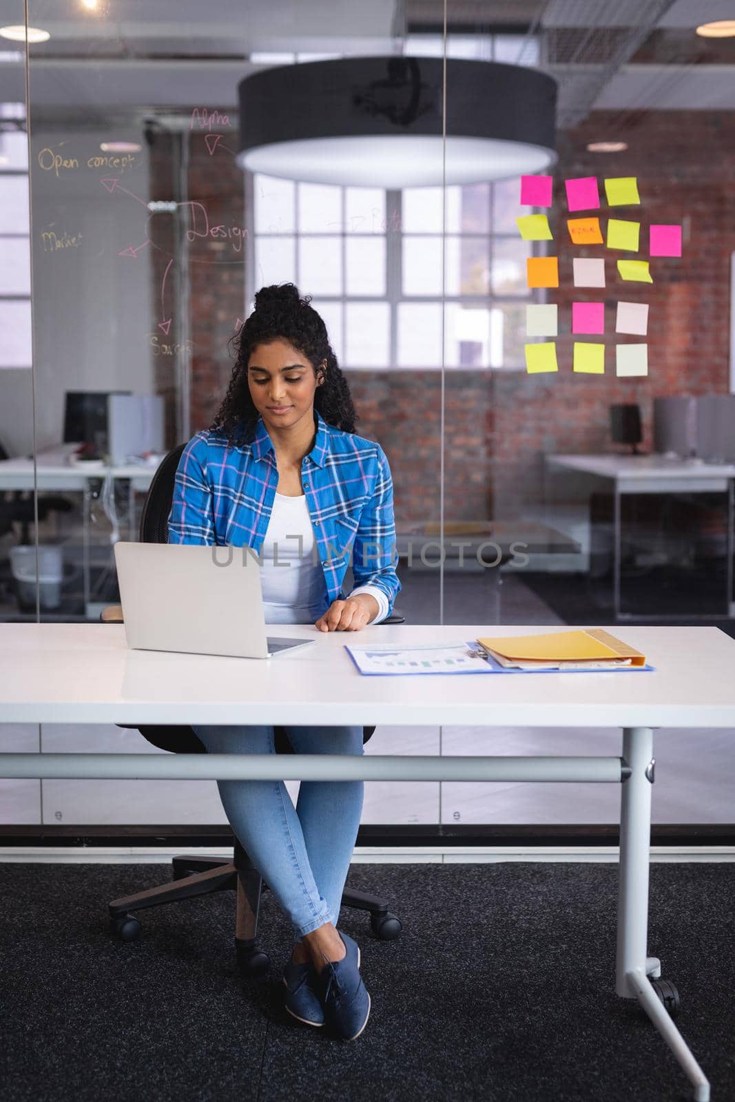 Mixed race businesswoman sitting in front of laptop with sticky memos on wall in background. independent creative design business.