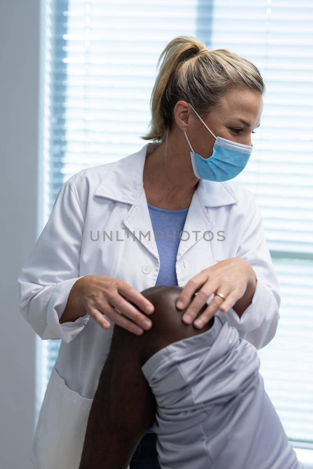 Caucasian female physiotherapist wearing face mask palpating knee of african american male patient. medicine, health and healthcare services during coronavirus covid 19 pandemic.