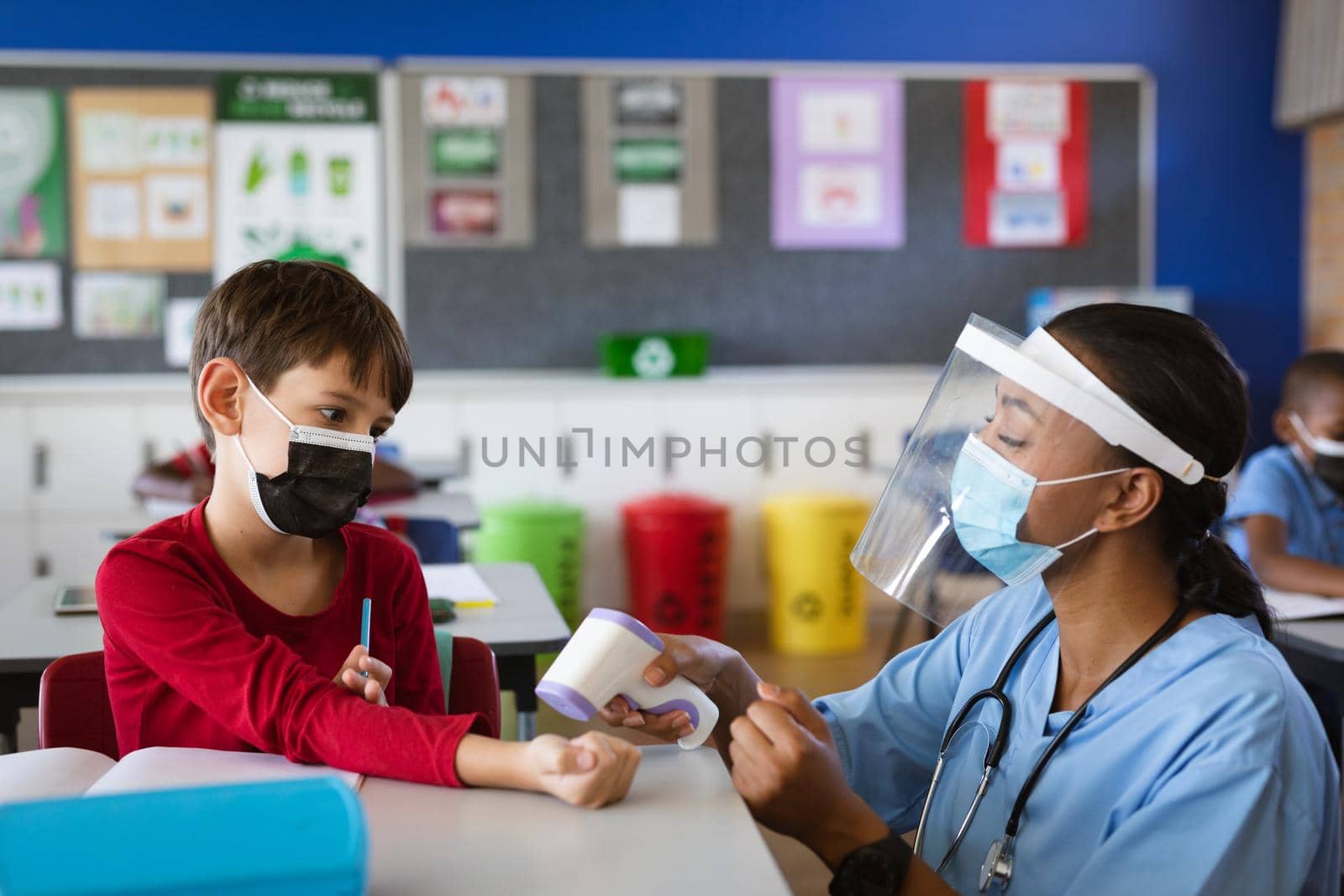 Female health worker wearing face shield measuring temperature of a boy at elementary school. education back to school health safety during covid19 coronavirus pandemic.