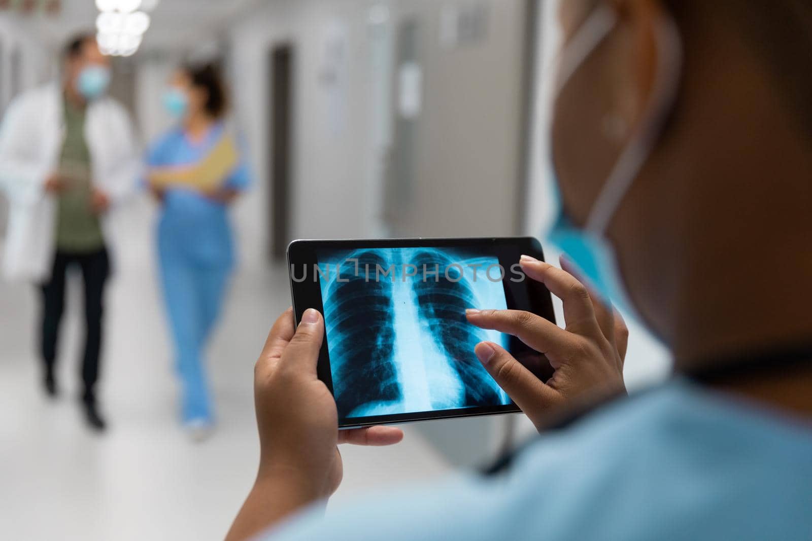 Mixed race doctor in corridor wearing face mask looking at lung x-ray on tablet. medicine, health and healthcare services during coronavirus covid 19 pandemic.