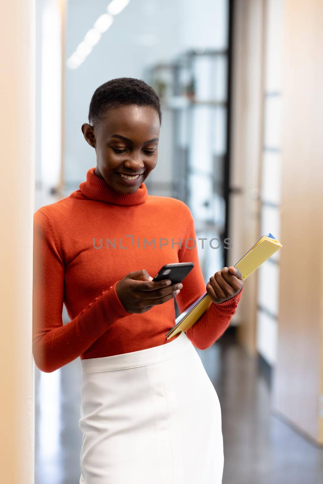Happy african american businesswoman standing in corridor using smartphone holding documents. business in a modern office.