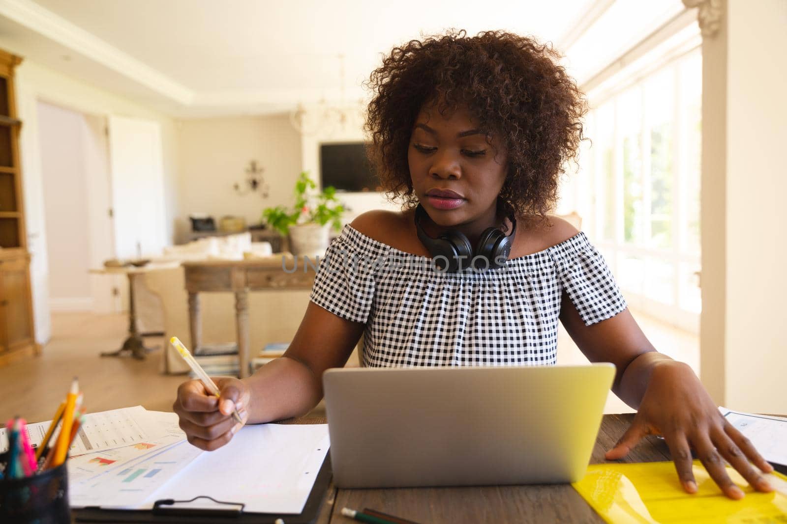African american woman wearing headphones, working in living room using laptop and making notes by Wavebreakmedia