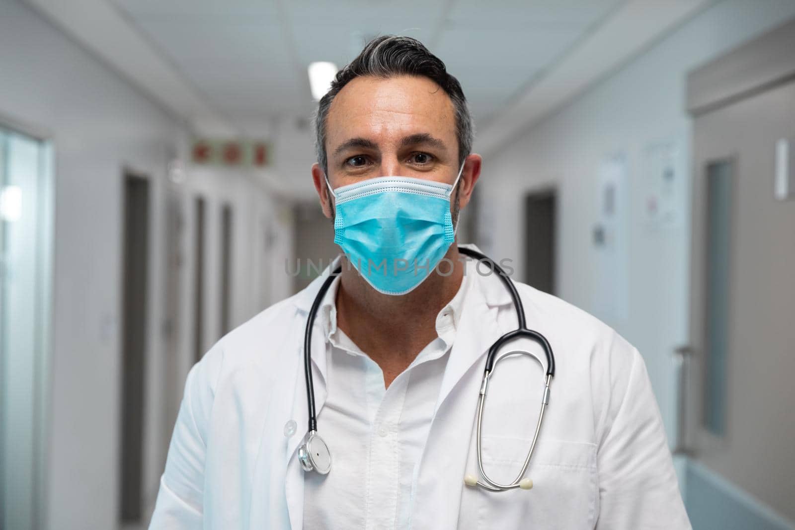 Portrait of mixed race male doctor wearing face mask standing in hospital corridor. medicine, health and healthcare services during coronavirus covid 19 pandemic.